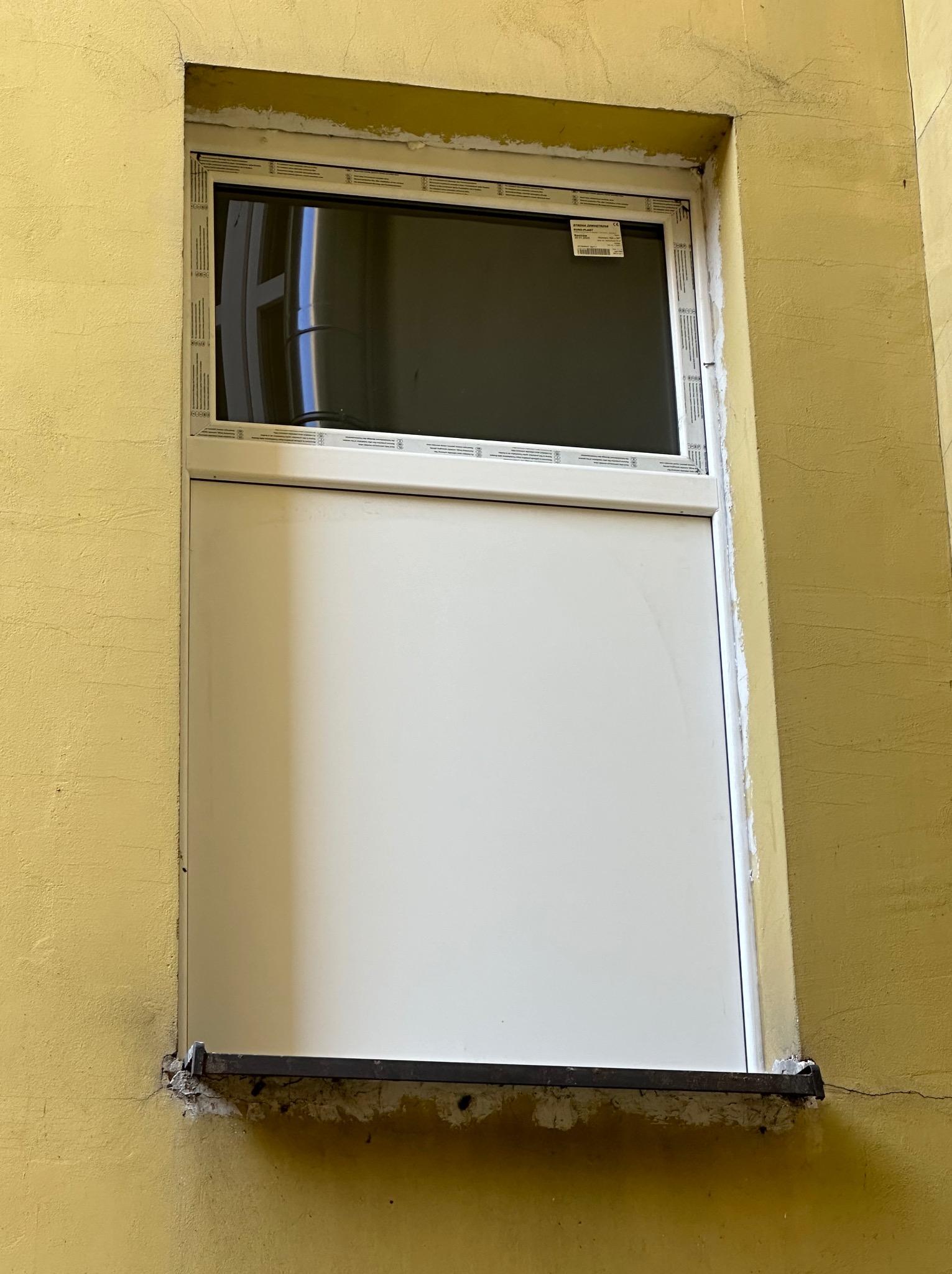 Białe okno PCV z szybą u góry i panelem na dole, zamontowane w żółtej ścianie budynku. Widoczne ślady prac montażowych i starej instalacji.