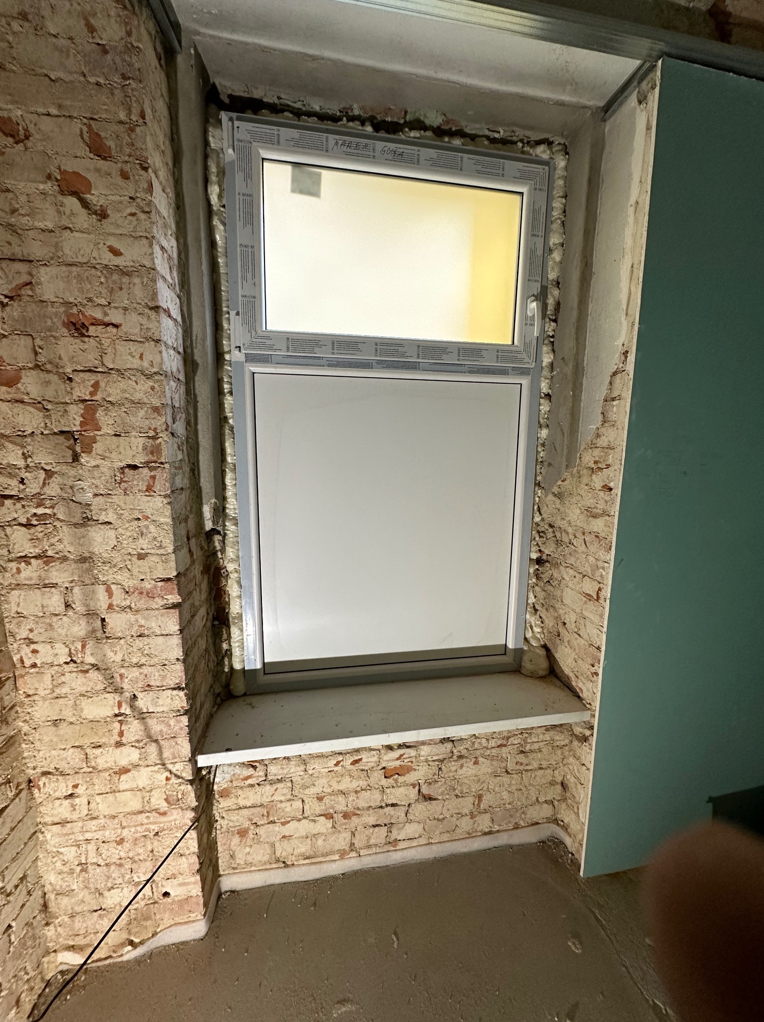 Wnętrze z nowym oknem PCV osadzonym w surowej, ceglanej wnęce. Widoczna pianka montażowa i częściowo wykończona ściana z płyty gipsowej.