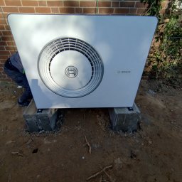 HYDROMAN Uladzislau Bliudnik - Biała pompa ciepła Bosch zamontowana na betonowych blokach przed ceglaną ścianą. Widoczny fragment pracownika instalującego urządzenie. Grunt ziemisty.