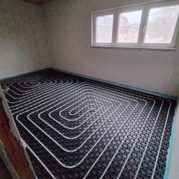 HYDROMAN Uladzislau Bliudnik - Białe rury ogrzewania podłogowego ułożone spiralnie na czarnej macie w surowym pomieszczeniu z oknem. Widok z góry, etap instalacji przed wylewką.