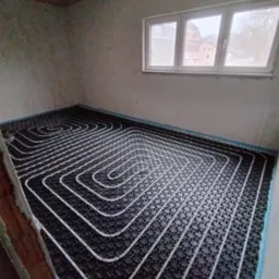 Białe rury ogrzewania podłogowego ułożone spiralnie na czarnej macie w surowym pomieszczeniu z oknem. Widok z góry, etap instalacji przed wylewką.