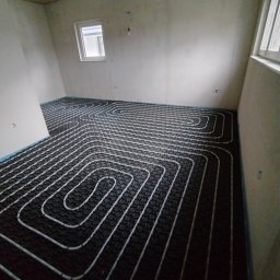HYDROMAN Uladzislau Bliudnik - Ułożone rury ogrzewania podłogowego na czarnej macie w surowym wnętrzu z oknem. Widok z góry ukazuje spiralny wzór instalacji.
