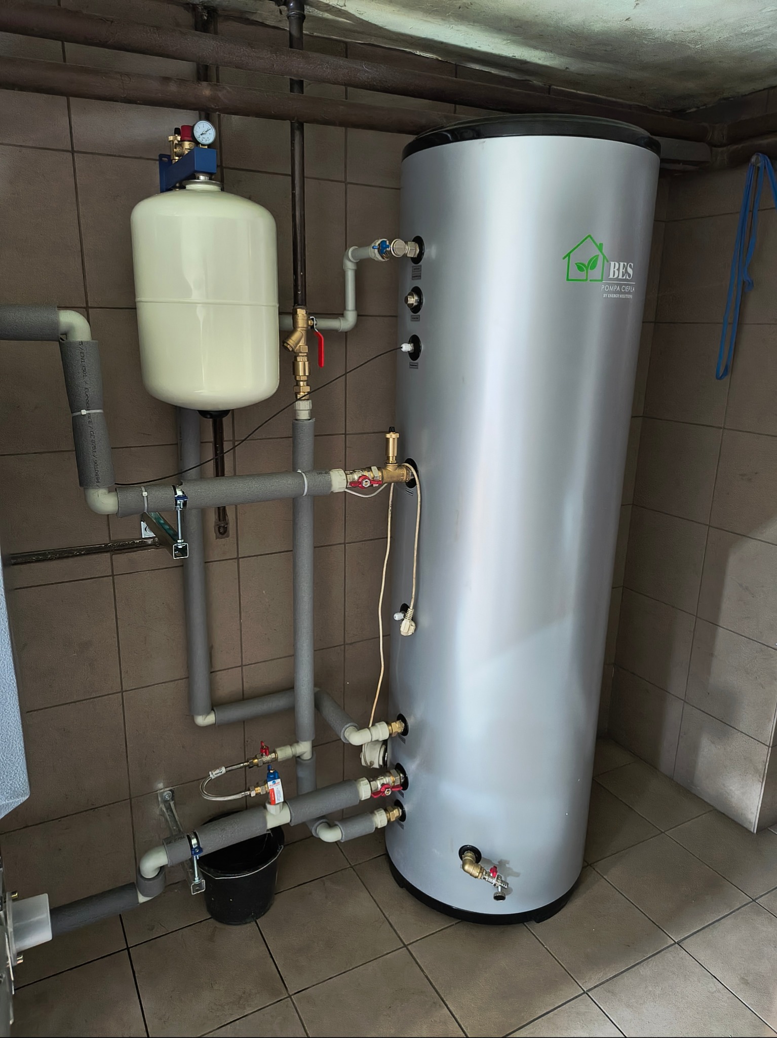 Zamontowany, srebrny zbiornik ciepłej wody użytkowej BES z podłączoną instalacją hydrauliczną w pomieszczeniu wyłożonym brązowymi płytkami.