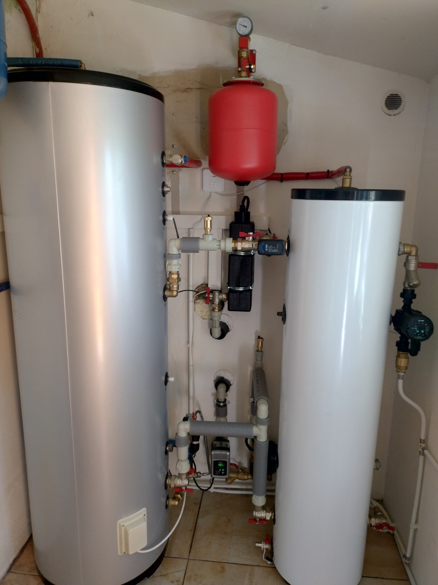 Dwa zbiorniki, srebrny i biały, z podłączeniami hydraulicznymi w pomieszczeniu technicznym. Czerwony zbiornik wyrównawczy z manometrem na górze.
