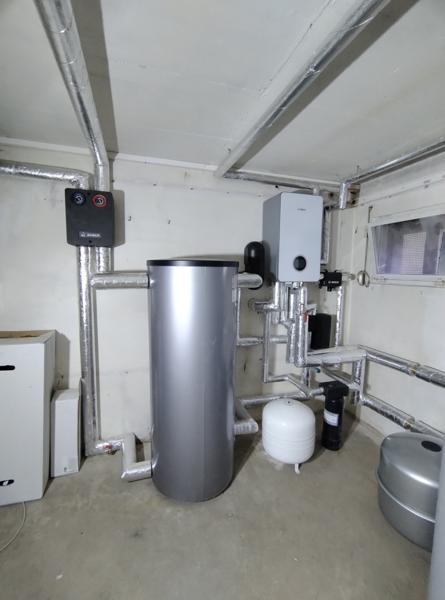 Srebrny zbiornik ciepłej wody użytkowej, biały piec gazowy Bosch i rury w izolacji aluminiowej w kotłowni. Kompaktowa instalacja grzewcza.