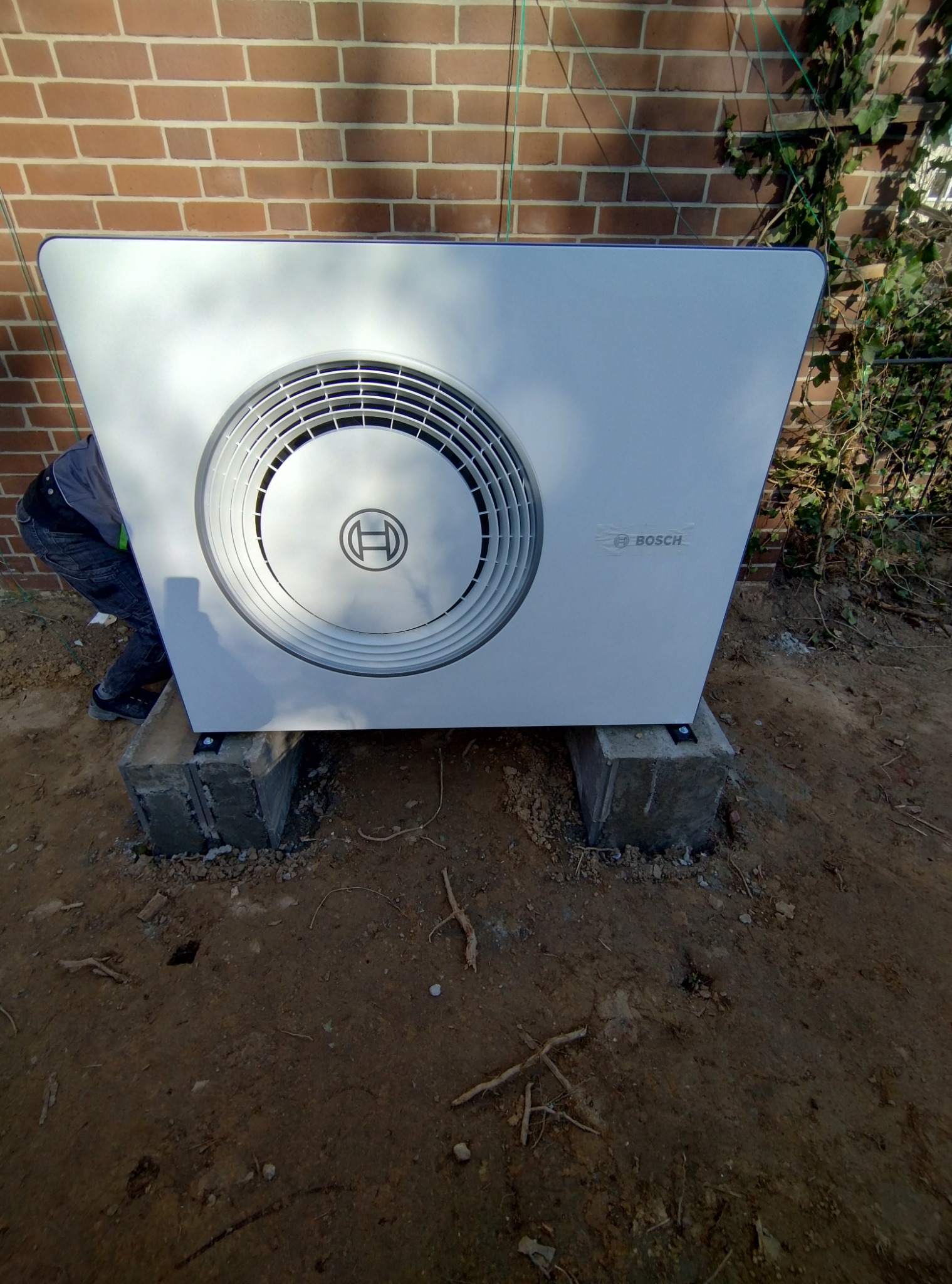 Biała pompa ciepła Bosch zamontowana na betonowych blokach przed ceglaną ścianą. Widoczny fragment pracownika instalującego urządzenie. Grunt ziemisty.