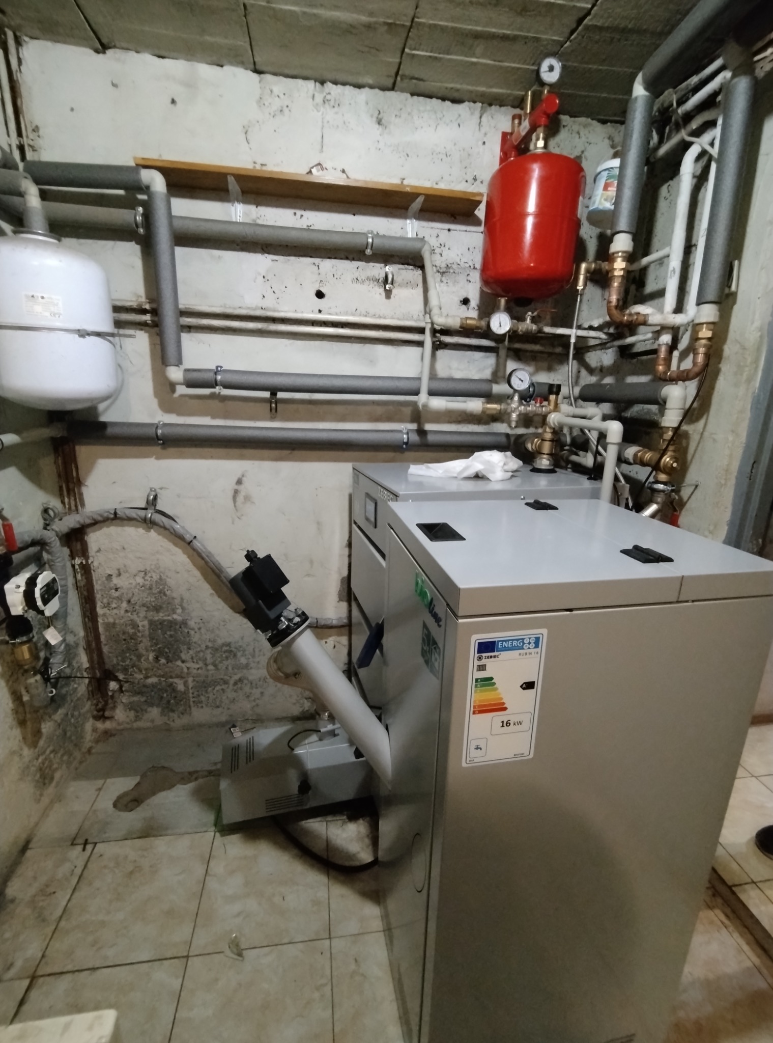 Zainstalowany piec gazowy Zebiec Rubin 16 kW w piwnicy, z widoczną instalacją hydrauliczną i grzewczą. Rury izolowane, manometry, zbiornik wyrównawczy.