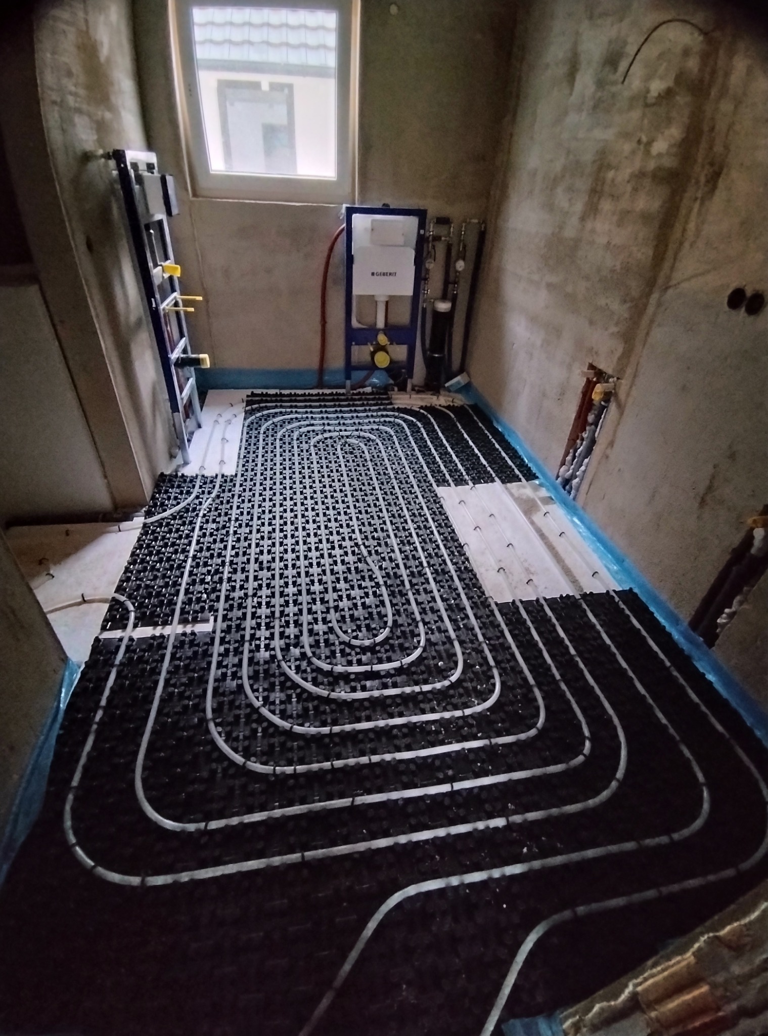 Wnętrze w surowym stanie z rozłożonym ogrzewaniem podłogowym na czarnej macie. Białe rurki tworzą regularny wzór. Widoczne okno i elementy instalacji.