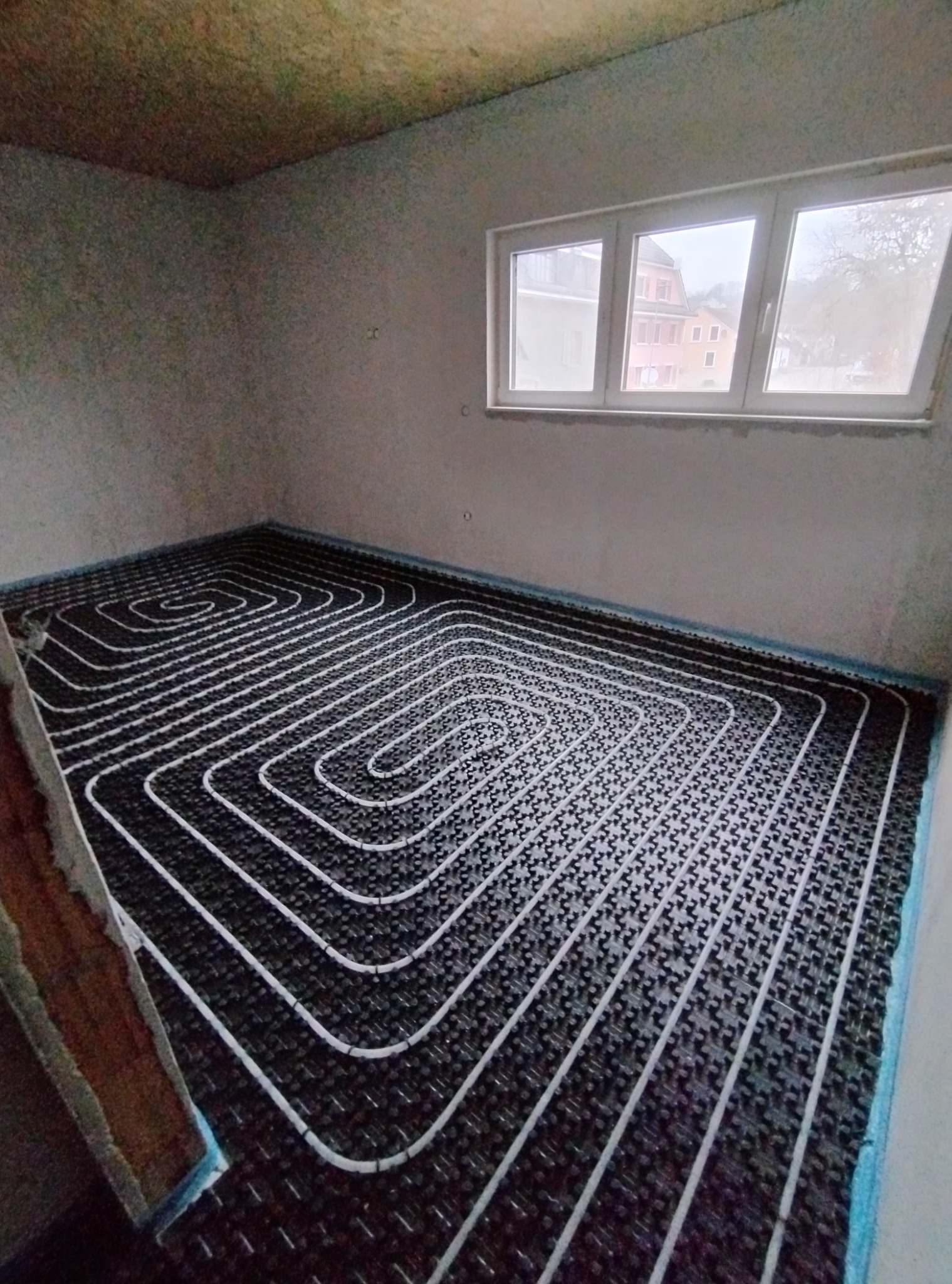 Białe rury ogrzewania podłogowego ułożone spiralnie na czarnej macie w surowym pomieszczeniu z oknem. Widok z góry, etap instalacji przed wylewką.