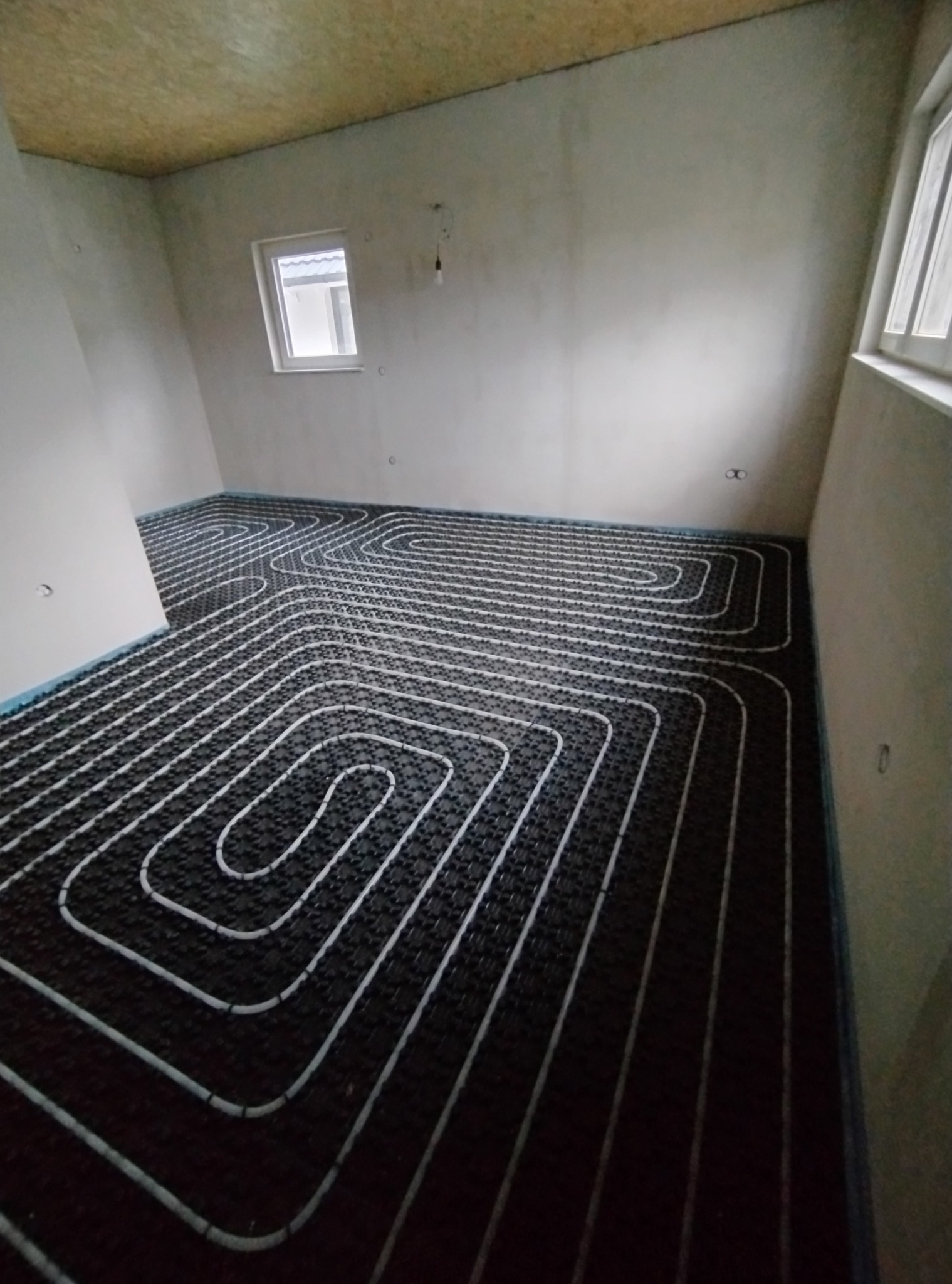 Ułożone rury ogrzewania podłogowego na czarnej macie w surowym wnętrzu z oknem. Widok z góry ukazuje spiralny wzór instalacji.