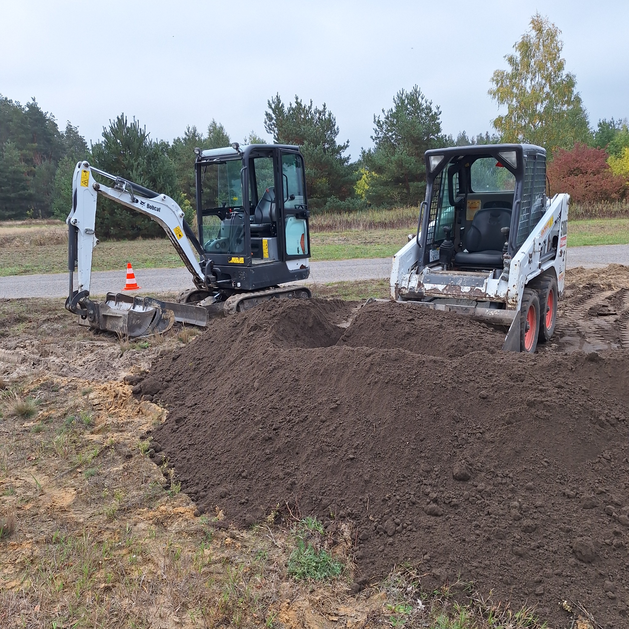 Dwa białe mini-ładowarki Bobcat na ziemi, z pryzmą ciemnej ziemi między nimi, w tle las i szare niebo. Jeden z nich ma pomarańczowy pachołek.