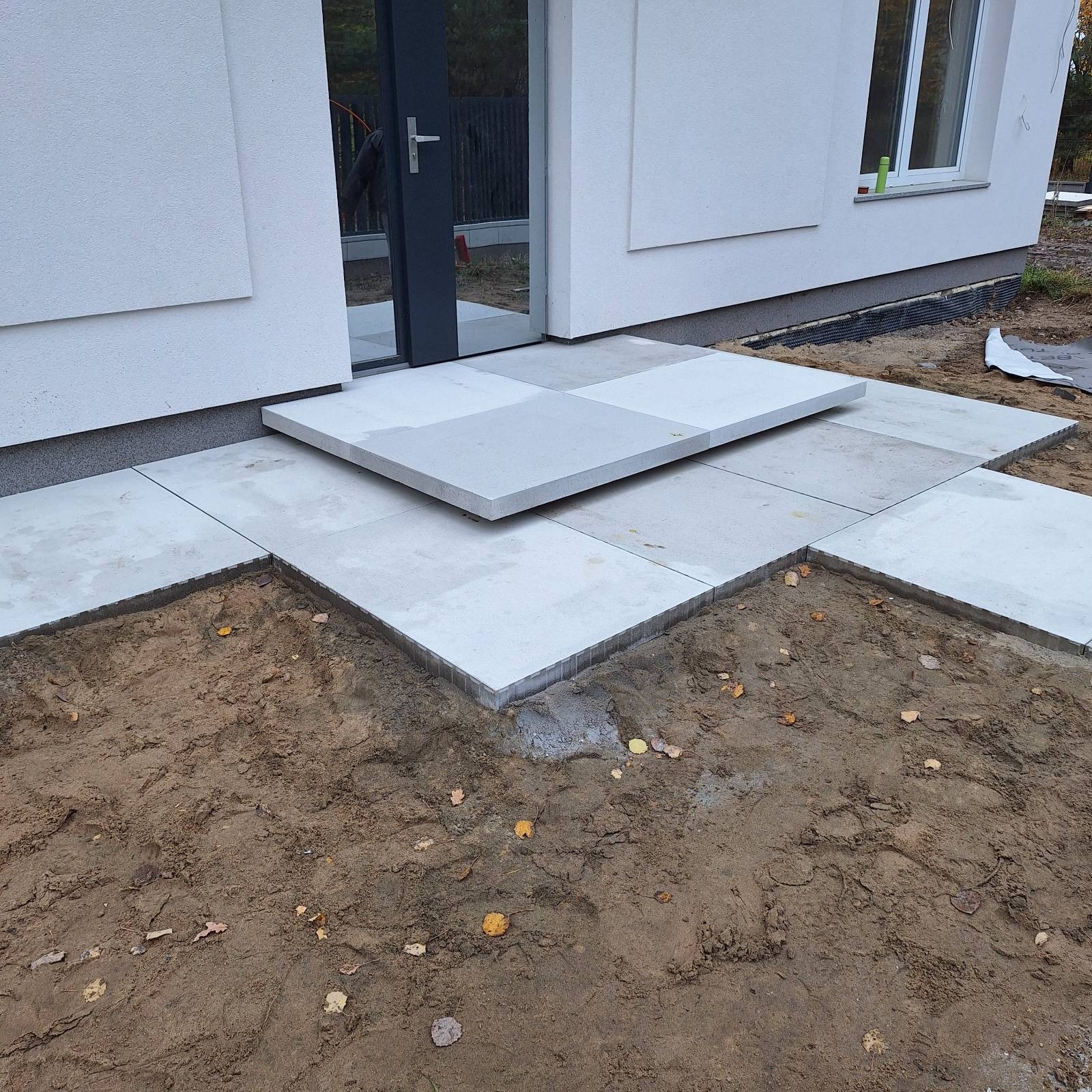 Nowoczesne, jasnoszare płyty chodnikowe ułożone przed wejściem do domu, z widoczną ziemią i jesiennymi liśćmi. Minimalistyczny design.