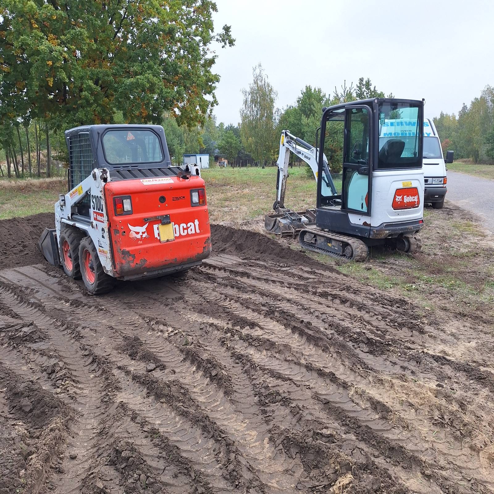 Dwa minikoparki Bobcat na polu z przygotowaną glebą, widoczne ślady opon. W tle drzewa i budynek. Prace ziemne w ogrodzie.