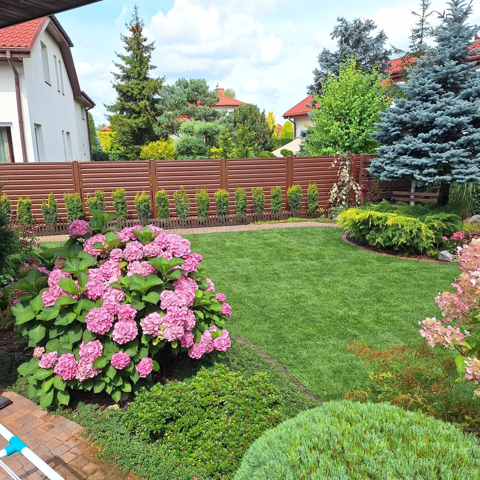 Zadbany ogród z równo przystrzyżonym trawnikiem, rabatami kwiatowymi i ozdobnymi krzewami, otoczony brązowym płotem. W tle widoczny dom z czerwonym dachem.
