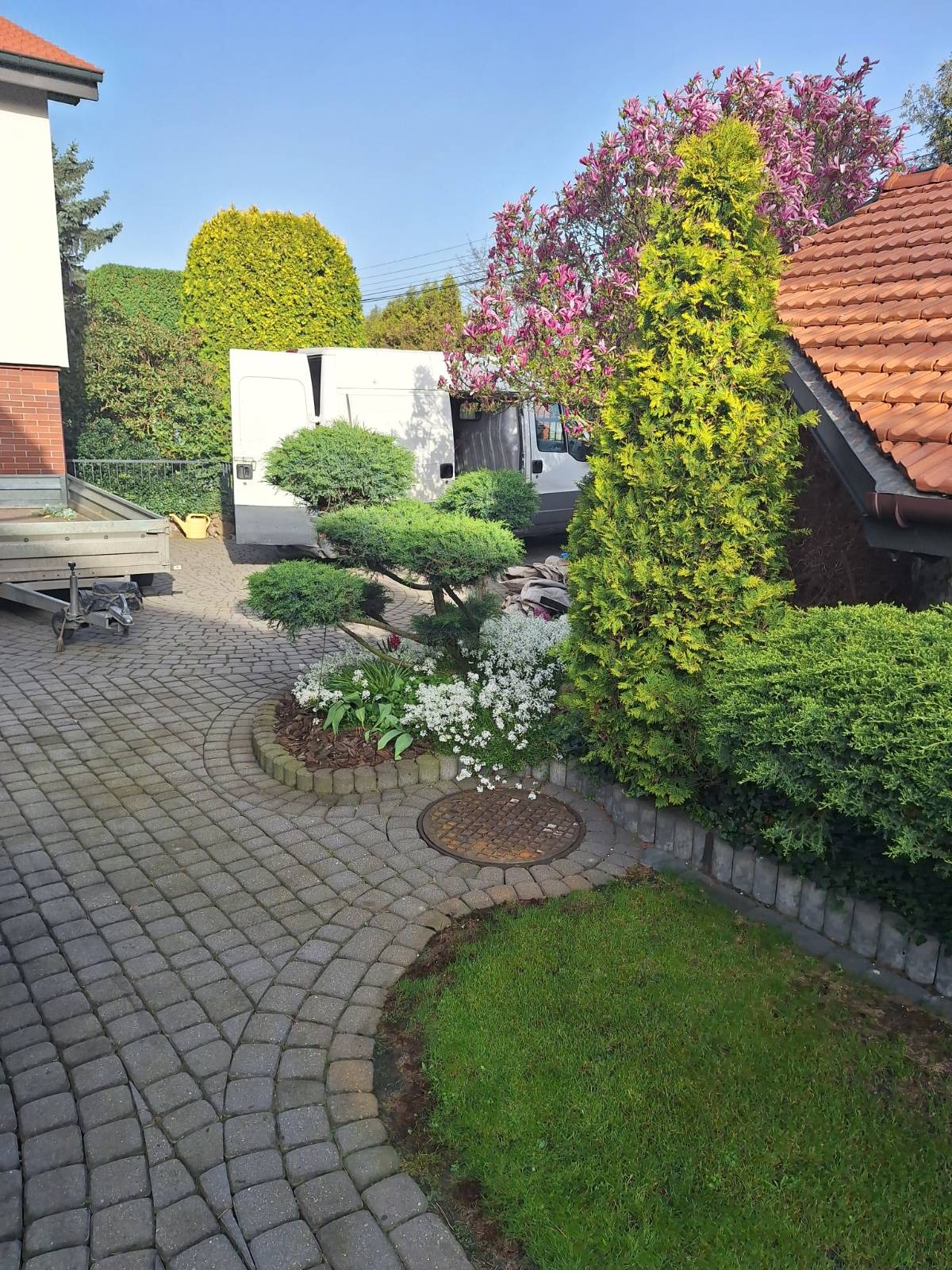 Uformowany ogród z krzewami bonsai i kwitnącą magnolią w tle, obok domu z czerwoną dachówką i zaparkowanym busem. Brukowany podjazd.