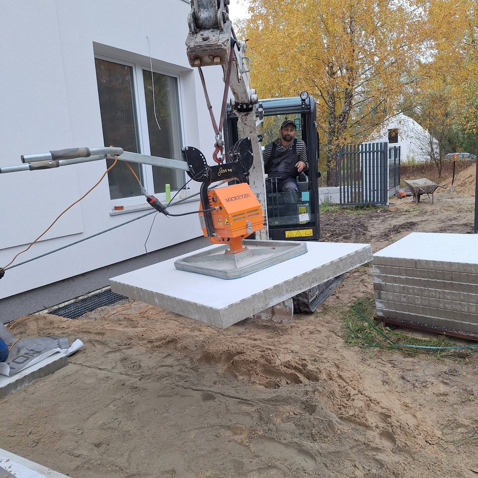 Operator mini-koparki układa betonowe płyty fundamentowe przy białym budynku. Widoczne narzędzia i przygotowany teren. Jesienne drzewa w tle.