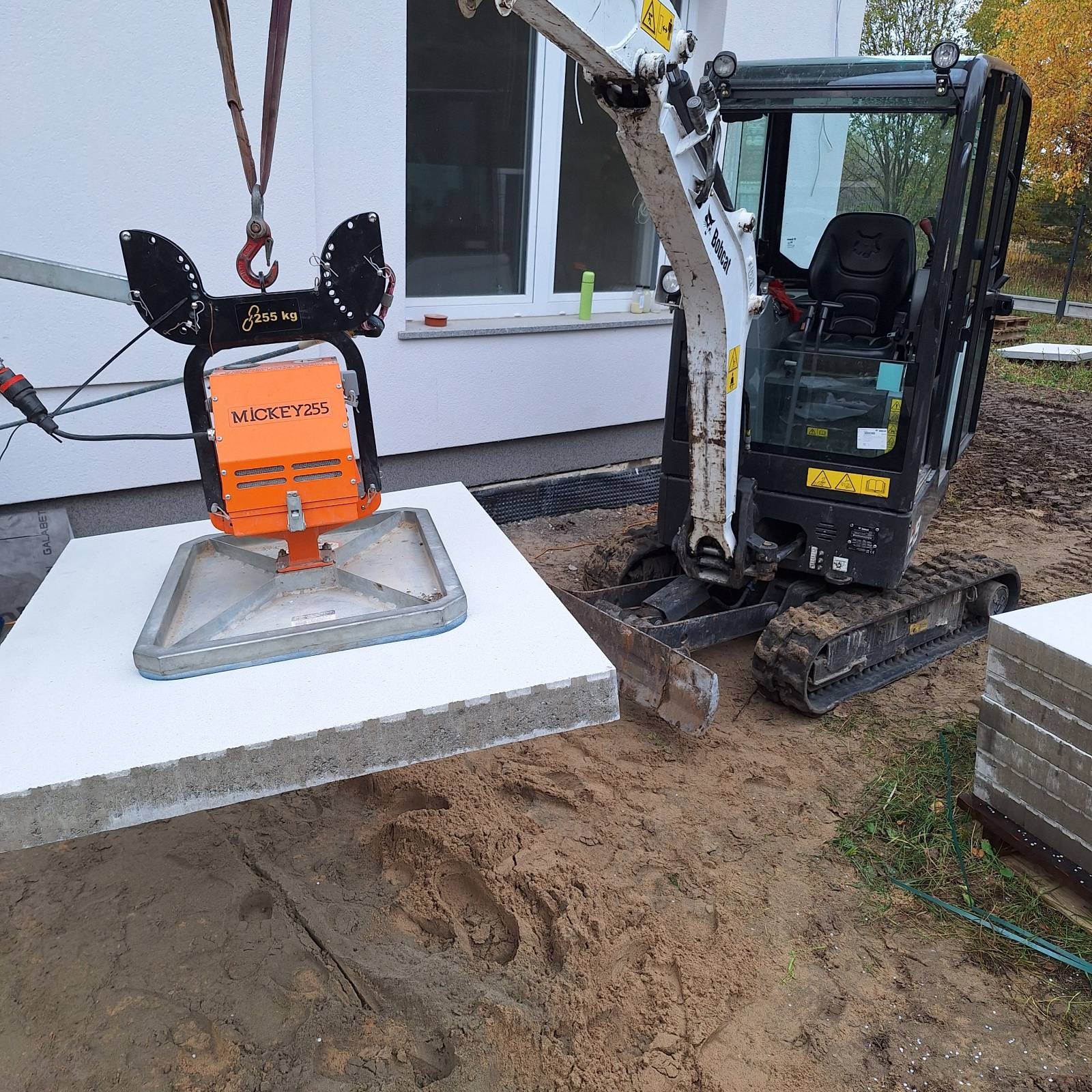 Mini koparka Bobcat podnosi płytę chodnikową za pomocą specjalistycznego chwytaka. Prace ziemne, przygotowanie terenu pod budowę.
