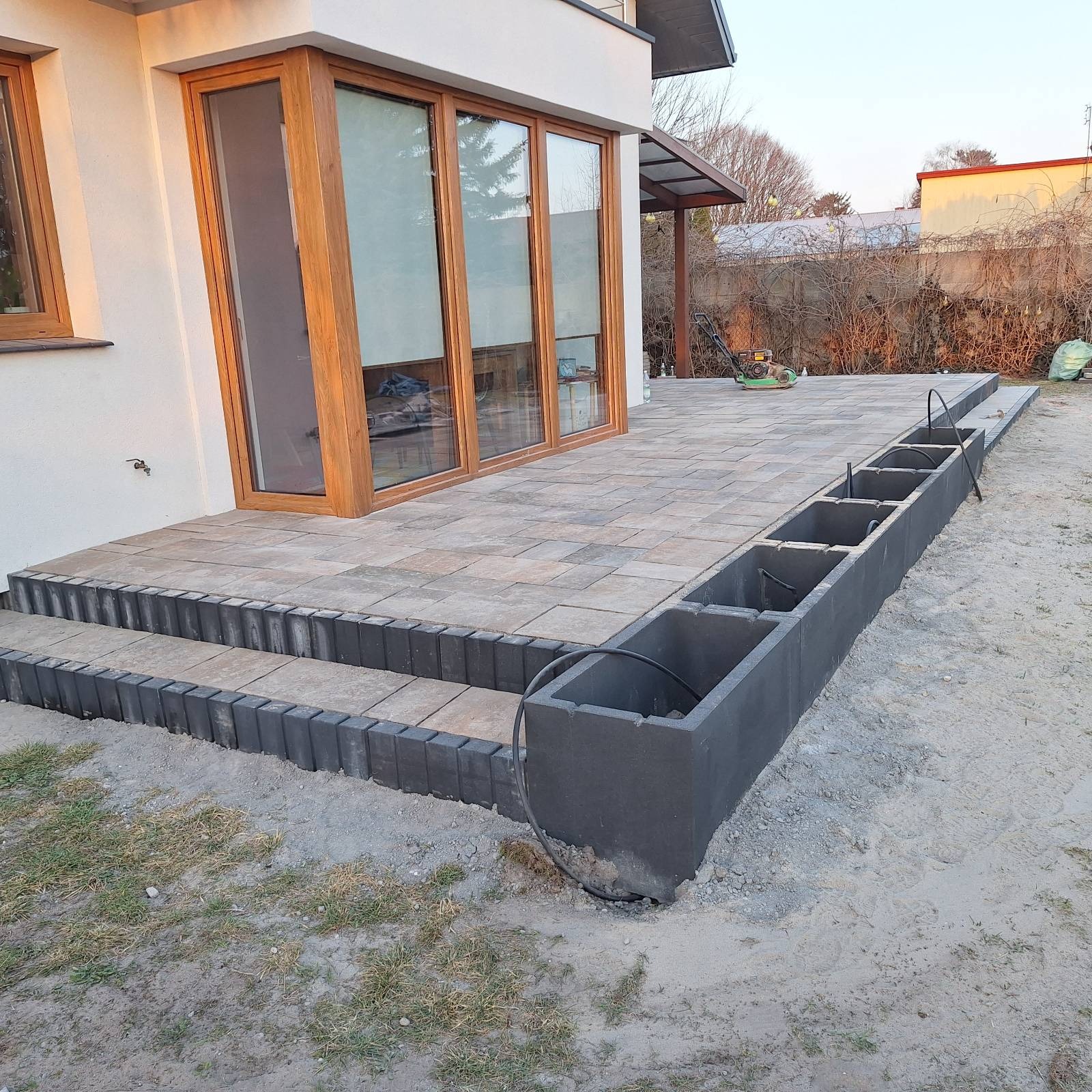 Wykończony taras z kostki brukowej z obrzeżami i schodami, widok na dom z drewnianą stolarką okienną. W tle trawnik i krzewy.