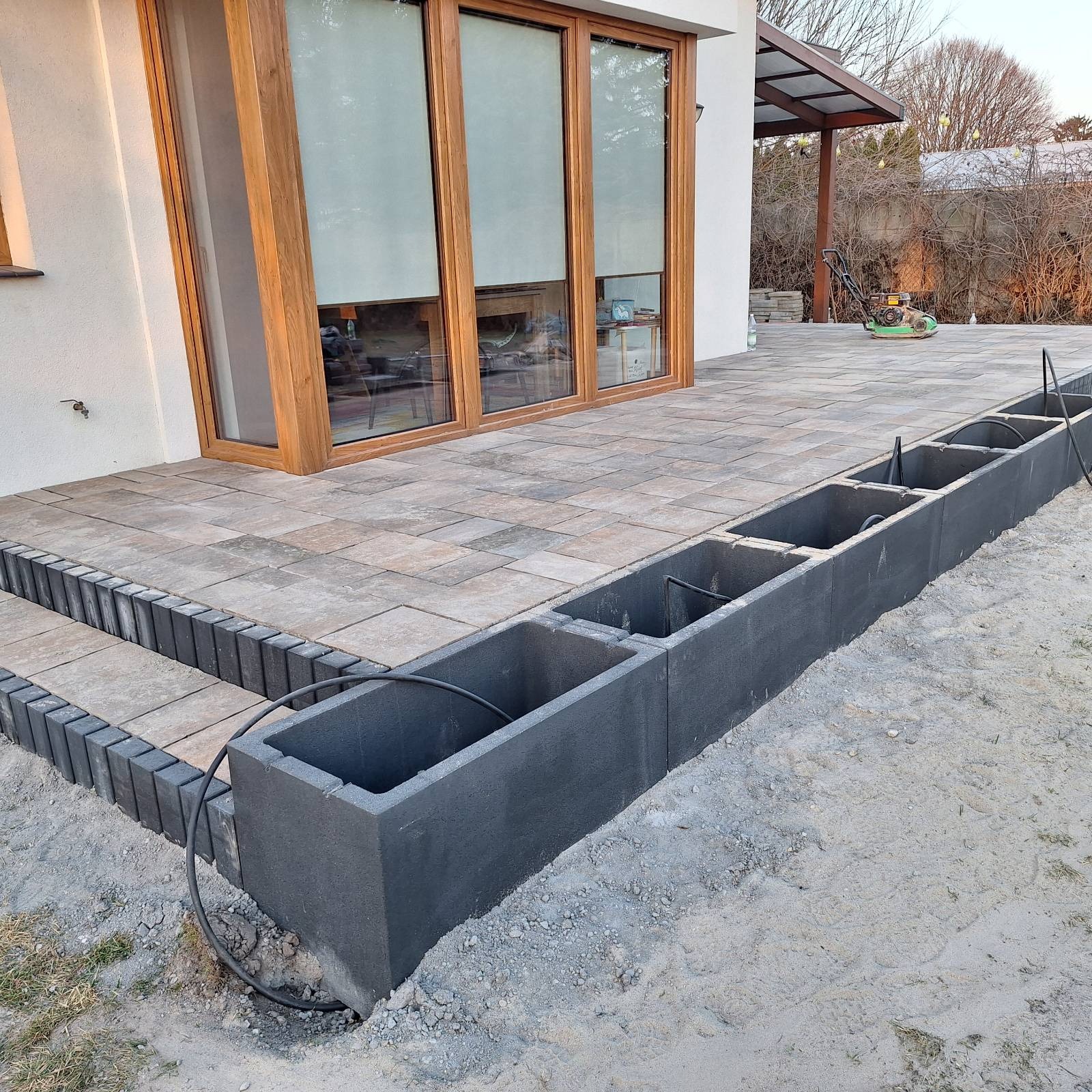 Układanie kostki brukowej na tarasie z przygotowanymi donicami z betonu. W tle widoczna maszyna do ubijania gruntu i okno tarasowe.
