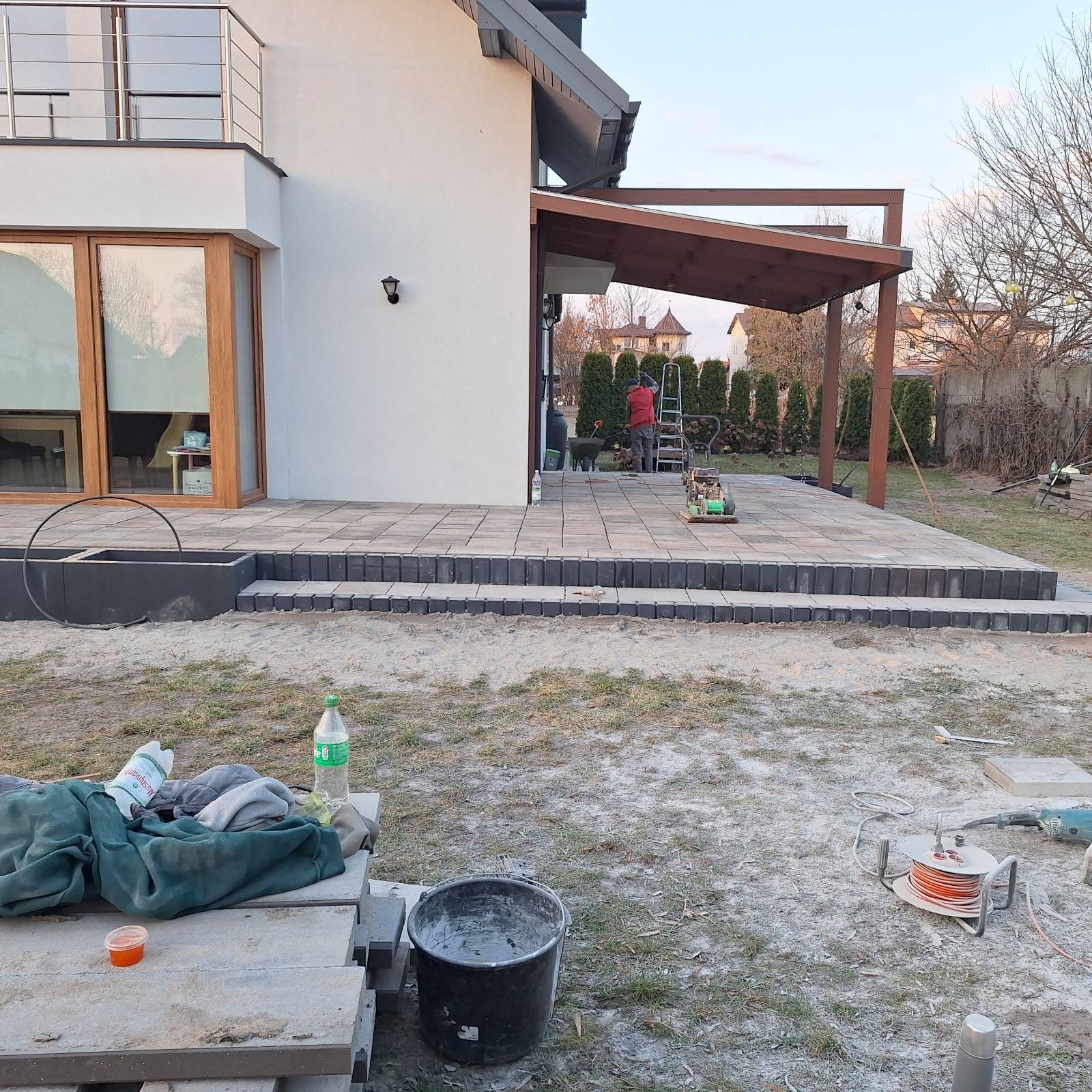 Układanie kostki brukowej na tarasie domu jednorodzinnego. Widoczny pracownik na drabinie, zagęszczarka i narzędzia. Częściowo wykończony taras z pergolą.