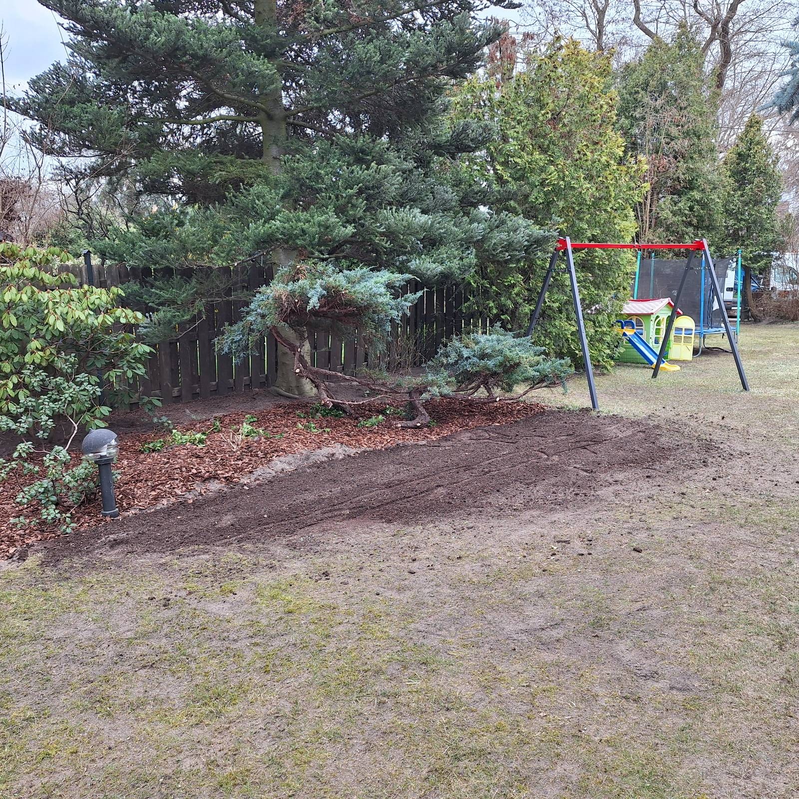 Ogród po przekopaniu ziemi pod nasadzenia, z korą wokół drzewa i elementami placu zabaw w tle. Widoczne ślady po pracy glebogryzarki.