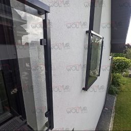 WOLMAR Edyta Wolna - Szklana balustrada balkonowa z czarnymi, metalowymi ramami i mocowaniami, widoczna z boku na tle białej ściany budynku i zieleni ogrodu. Minimalistyczny design.