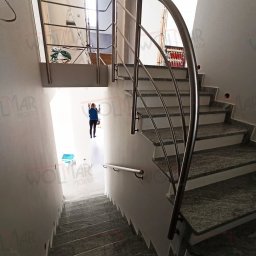 WOLMAR Edyta Wolna - Spiralne schody z szarego kamienia z balustradą ze stali nierdzewnej w Mokrej. Widok z góry na dół, osoba na dole schodów. Jasne wnętrze.