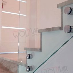 WOLMAR Edyta Wolna - Szklana balustrada schodowa z marmurowymi stopniami i srebrnymi mocowaniami. Widoczne logo firmy na ścianie. Detal nowoczesnego wykończenia wnętrza.