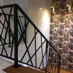 WOLMAR Edyta Wolna - Nowoczesna, czarna balustrada schodowa z geometrycznym wzorem na tle ściany w kwiaty i złotych lamp wiszących. Widok z dołu schodów, podkreślający unikalny design.