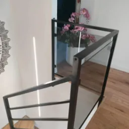 Nowoczesne schody z drewnianymi stopniami i czarną, metalową balustradą z elementami szklanymi, widziane z góry. Na ścianie dekoracyjny ornament.