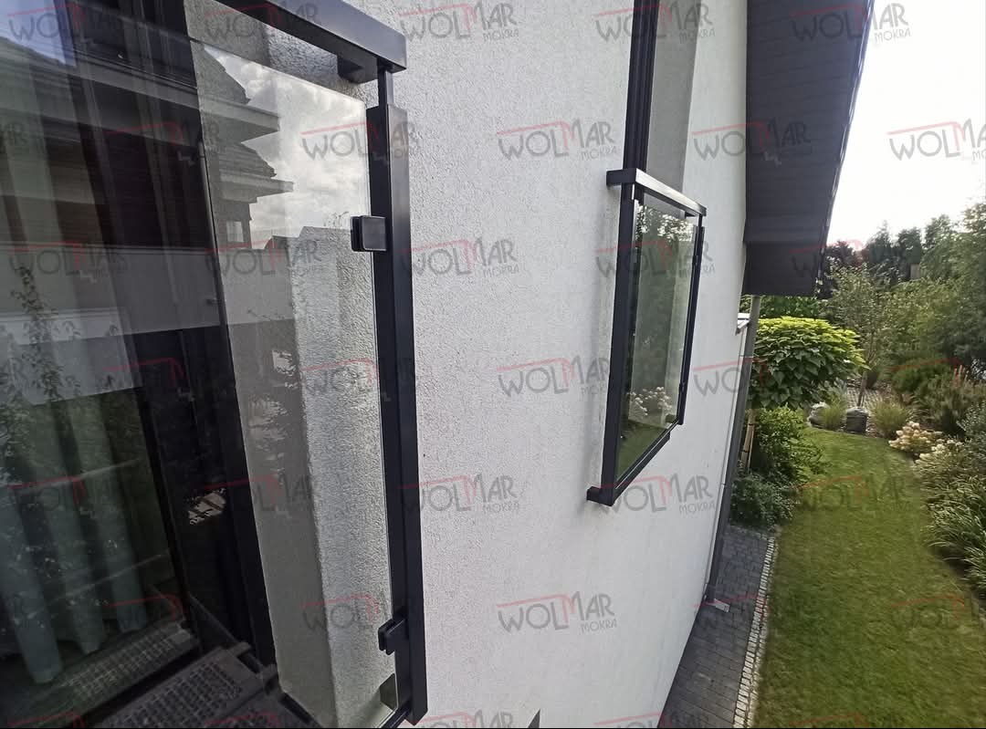 Szklana balustrada balkonowa z czarnymi, metalowymi ramami i mocowaniami, widoczna z boku na tle białej ściany budynku i zieleni ogrodu. Minimalistyczny design.