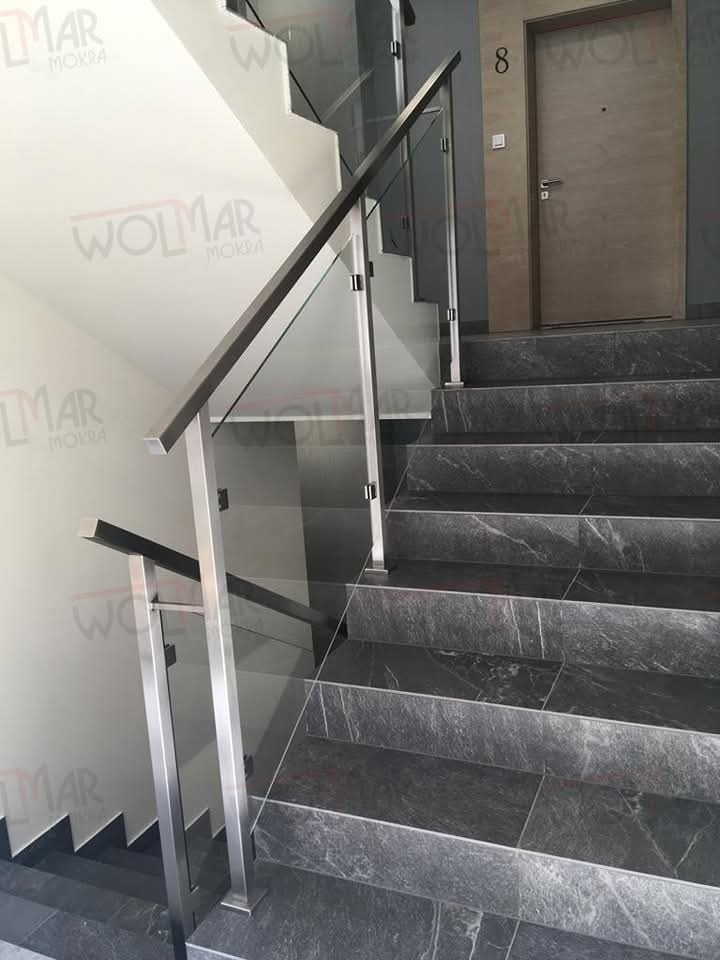 Nowoczesne schody z szarymi, marmurkowymi stopniami i balustradą ze stali nierdzewnej z wypełnieniem szklanym. Widoczne drzwi z numerem 8.