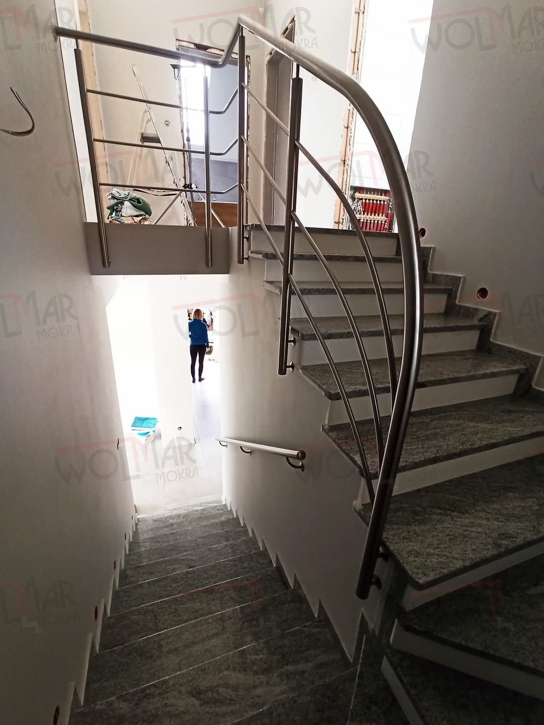 Spiralne schody z szarego kamienia z balustradą ze stali nierdzewnej w Mokrej. Widok z góry na dół, osoba na dole schodów. Jasne wnętrze.