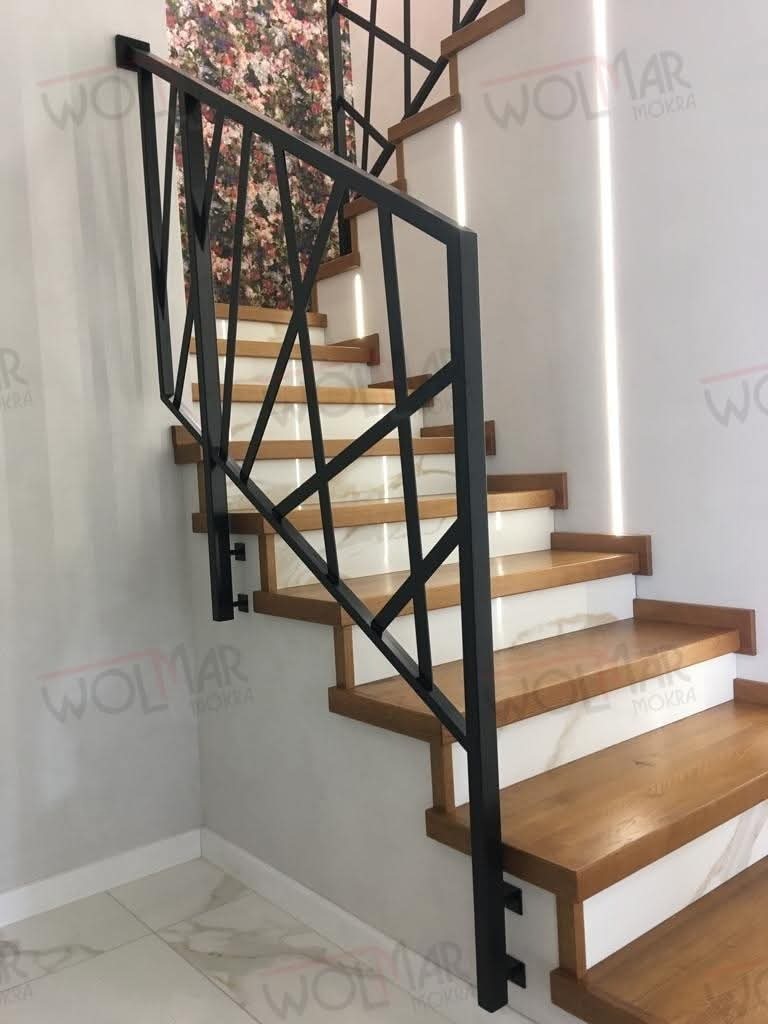 Nowoczesna, czarna poręcz schodowa z geometrycznym wzorem na drewnianych schodach z marmurowymi podstopnicami, oświetlona naturalnym światłem.