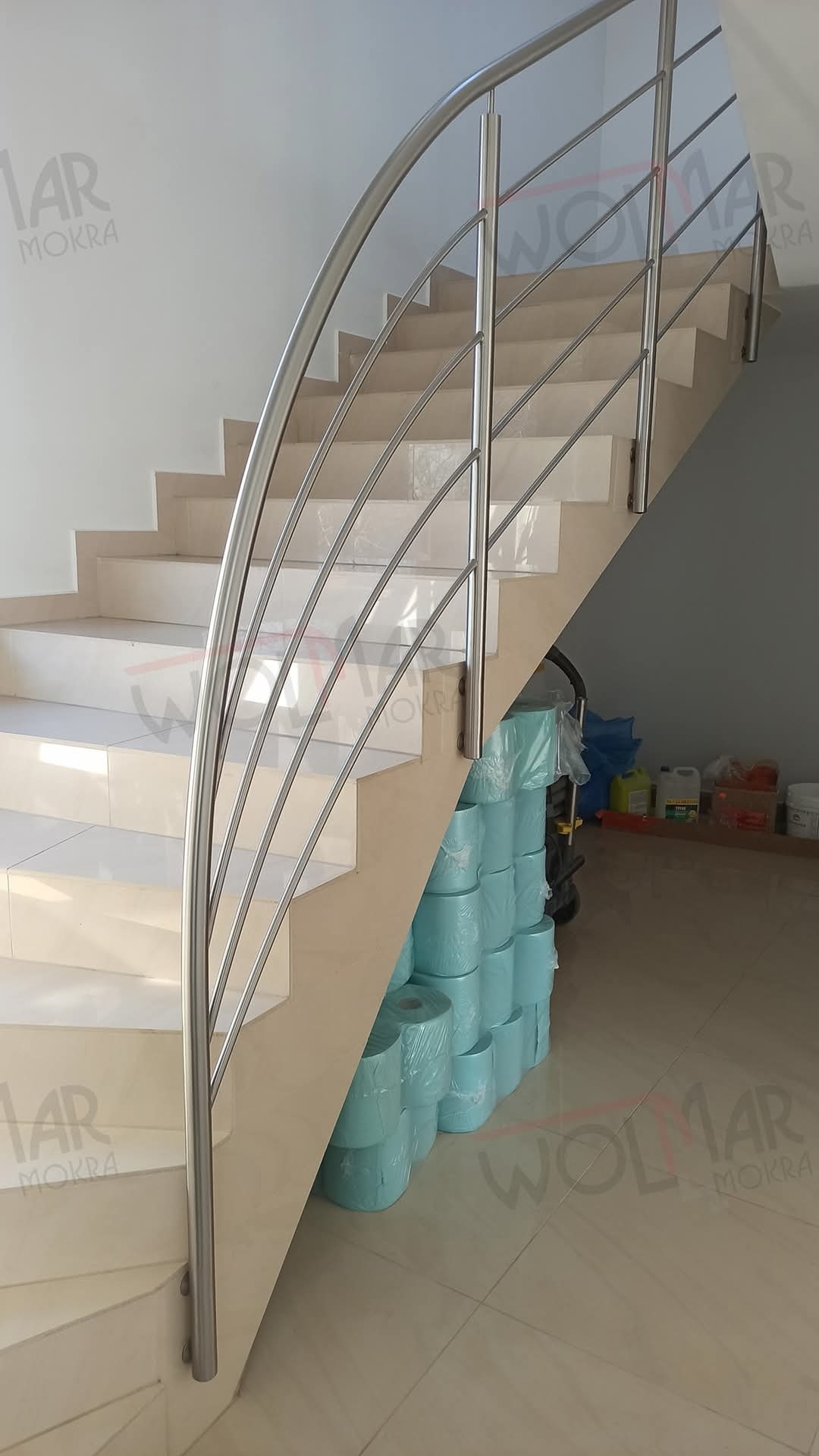 Zakrzywiona, metalowa balustrada ze stali nierdzewnej, zamontowana na jasnych, wyłożonych kafelkami schodach wewnętrznych. Widoczne materiały budowlane w tle.