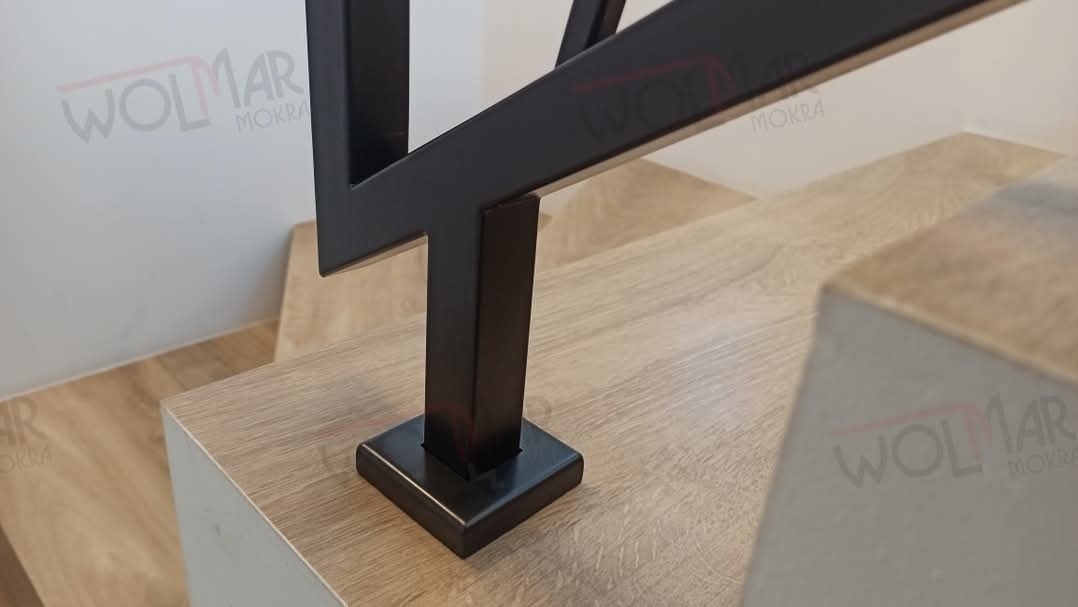 Fragment metalowej balustrady schodowej w kolorze czarnym, z geometrycznym wzorem, zamocowanej na drewnopodobnych stopniach. Widoczne detale mocowania.