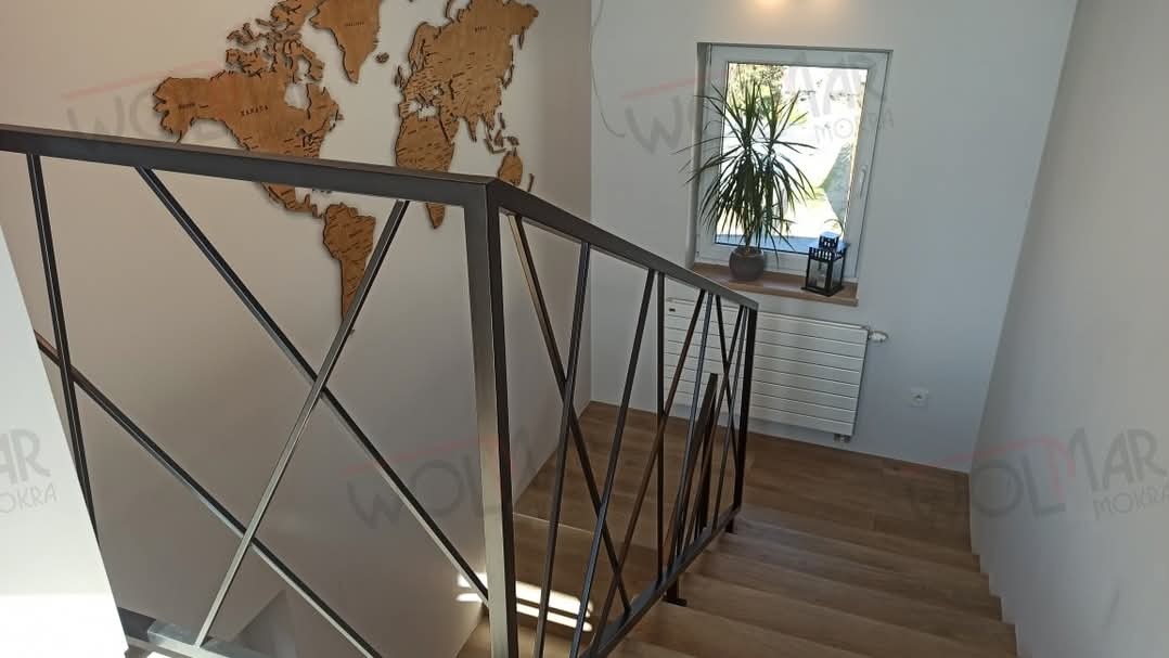 Metalowa balustrada schodowa z geometrycznym wzorem krzyżujących się prętów na tle drewnianej mapy świata i okna z rośliną.
