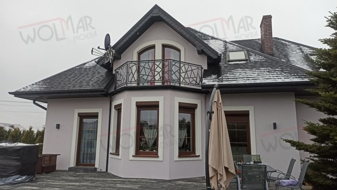 Szara elewacja domu z balkonem i geometryczną, czarną balustradą. Widoczne okna z brązowymi ramami, dach pokryty gontem bitumicznym i częściowo zasłonięty parasol.