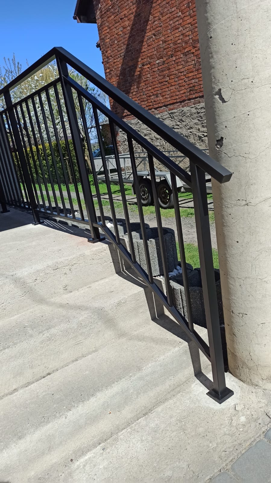 Czarna, metalowa poręcz schodowa z pionowymi szczeblami, zamontowana przy betonowych schodach zewnętrznych, na tle ceglanego budynku i przyczepy.