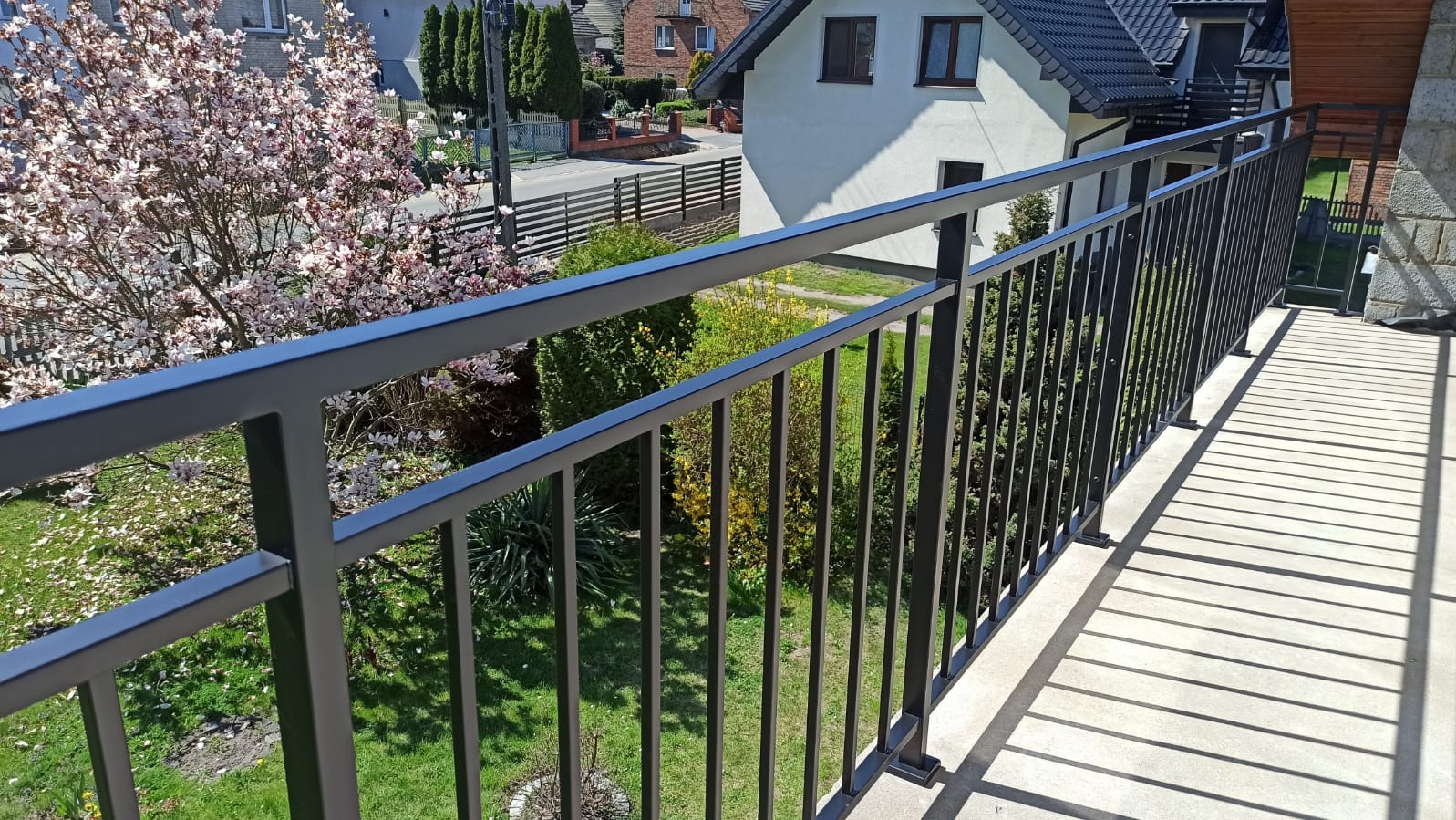 Nowoczesna, czarna, metalowa balustrada balkonowa z widokiem na ogród i kwitnące drzewo. Minimalistyczny design, proste linie, jasne światło słoneczne.