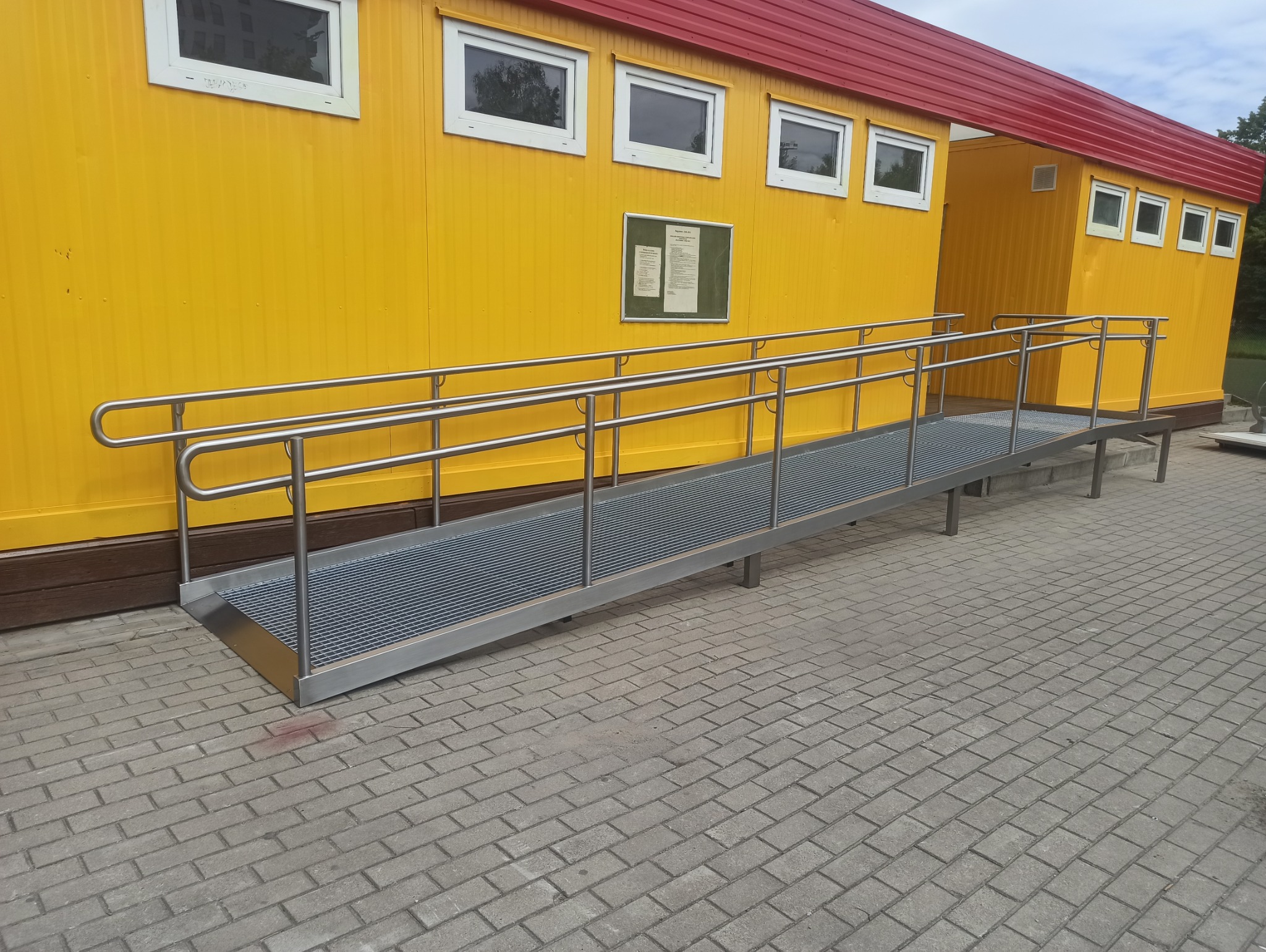 Rampa dla niepełnosprawnych z metalową kratą i poręczami, zamontowana przed żółtym budynkiem z czerwonym dachem, widok z boku na kostkę brukową.