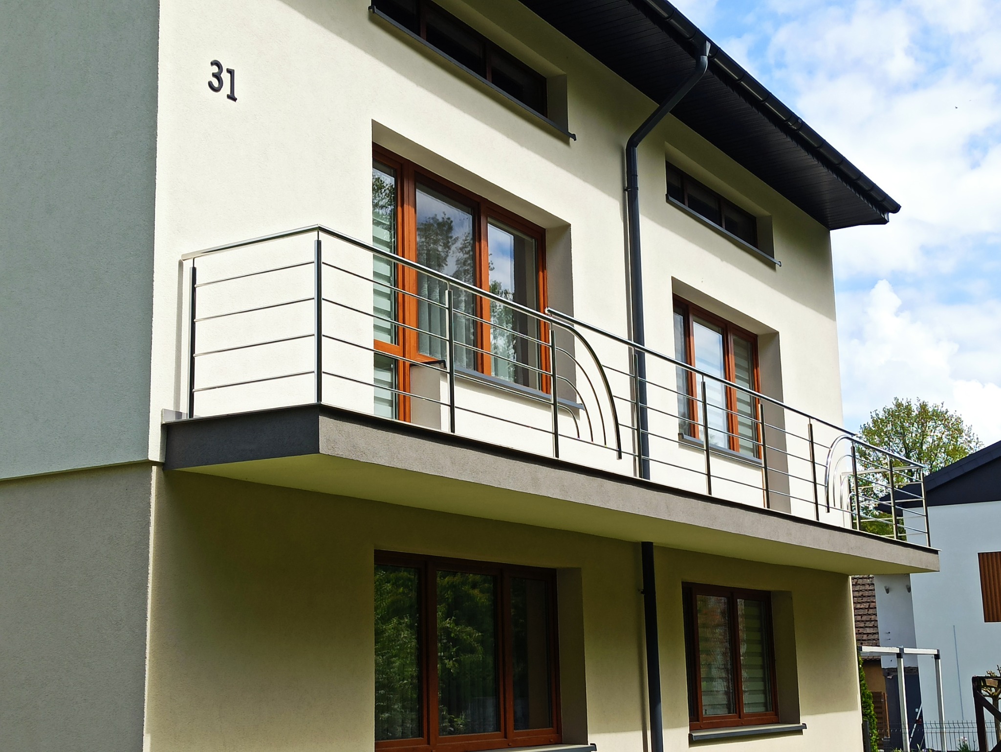 Balkon w nowoczesnym domu z chromowaną balustradą o minimalistycznym designie i elementami giętymi. Widoczne okna z drewnianą stolarką i numer domu '31'.