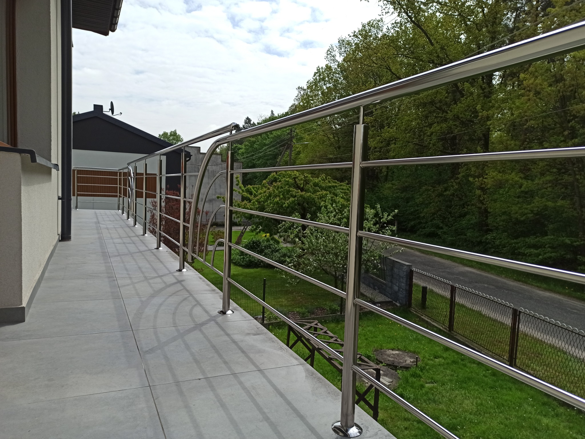 Błyszcząca, metalowa balustrada balkonowa z elementami dekoracyjnymi, na tle zieleni ogrodu i fragmentu ulicy. Nowoczesny design i precyzja wykonania.