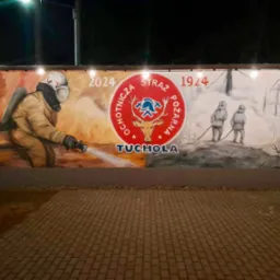 Mural przedstawiający strażaka w akcji gaszenia pożaru, obok logo OSP Tuchola z datami 1924 i 2024, namalowany na ścianie budynku.