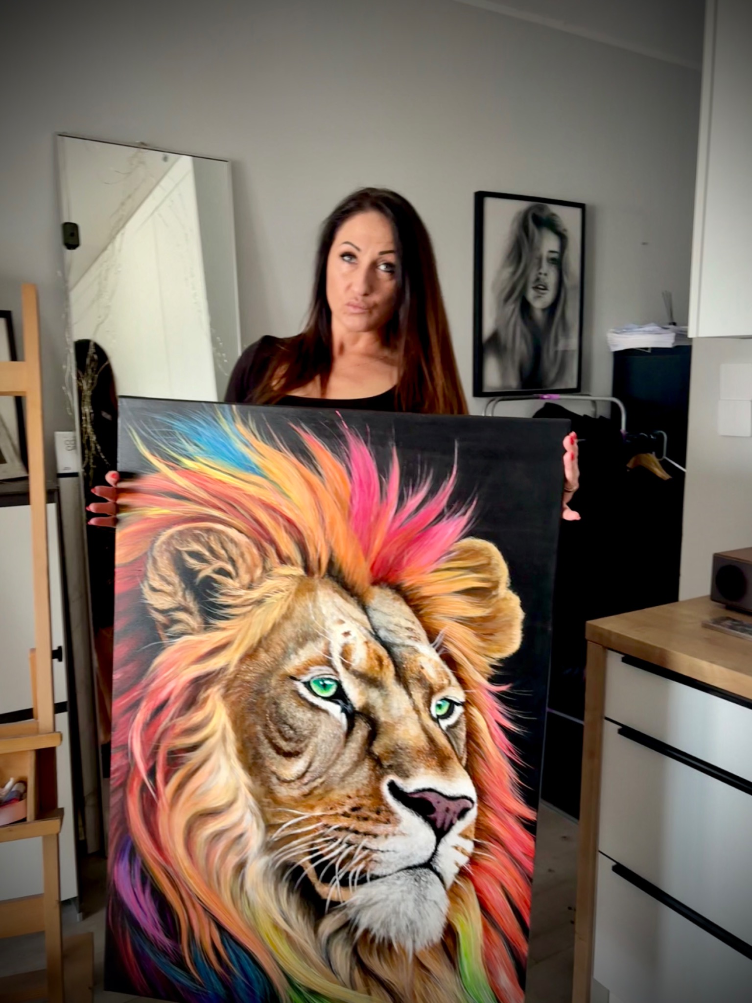 Artystka prezentuje ukończony, kolorowy obraz lwa na płótnie, trzymając go w pracowni. Widoczny stojak malarski i rama z portretem kobiety w tle.