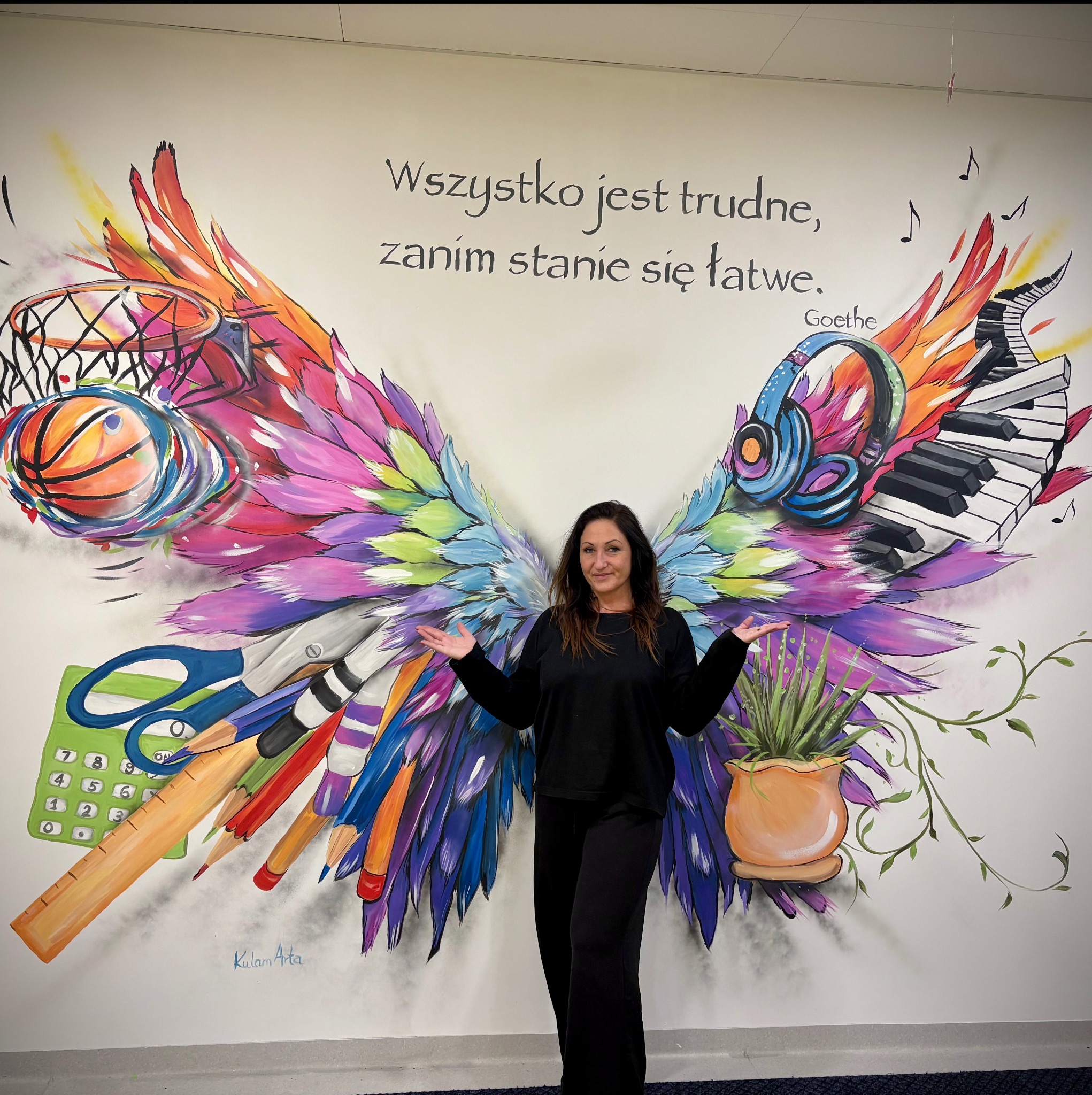 Kobieta na tle ściany z muralem skrzydeł, które tworzą kosz, słuchawki, pianino, przybory szkolne i doniczka z rośliną. Całość uzupełnia napis.
