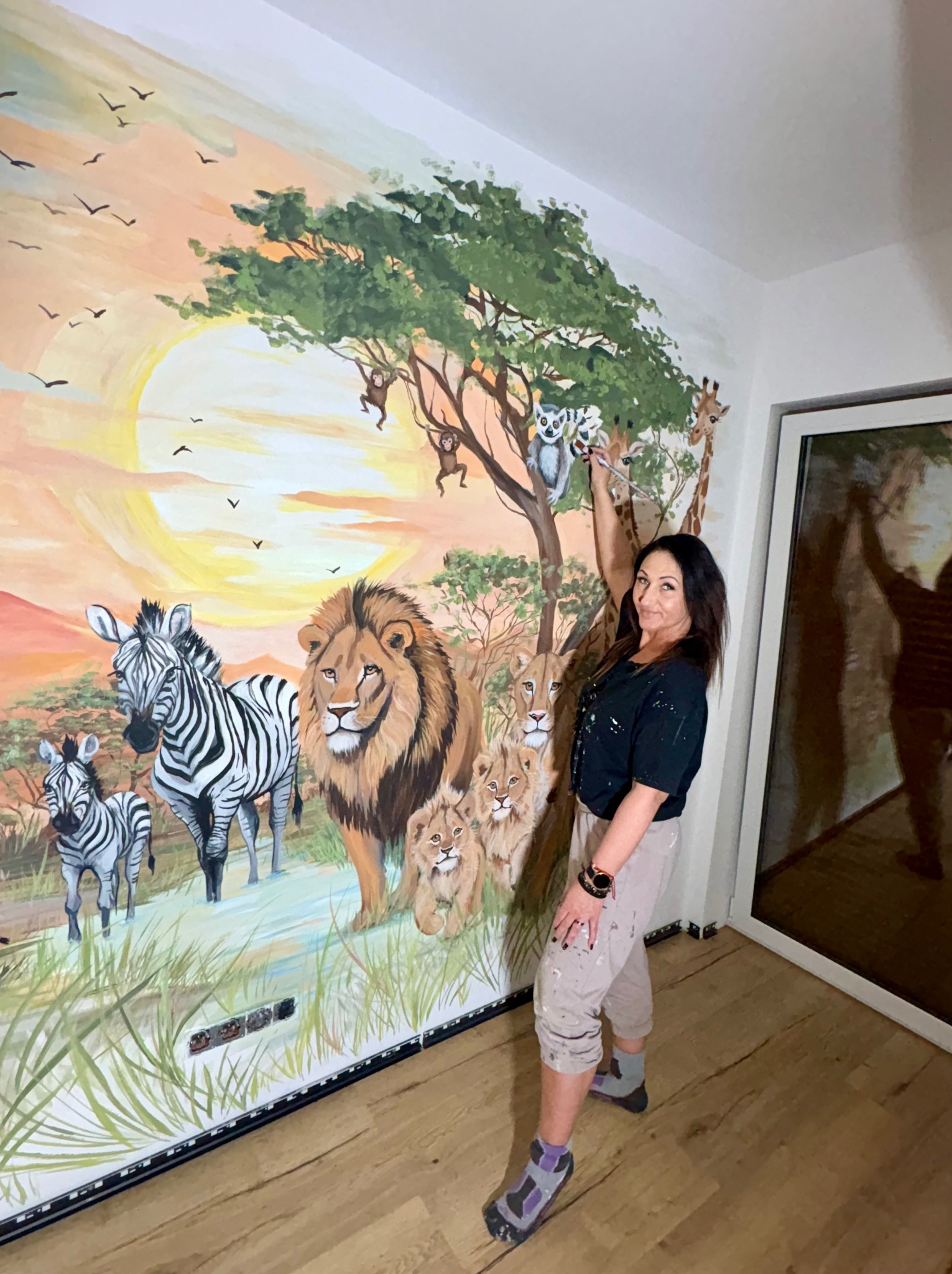Kobieta maluje na ścianie mural z motywem afrykańskiej sawanny: lwy, zebry, żyrafa, lemur i małpy na tle zachodzącego słońca. Widoczne ślady farby na ubraniu.