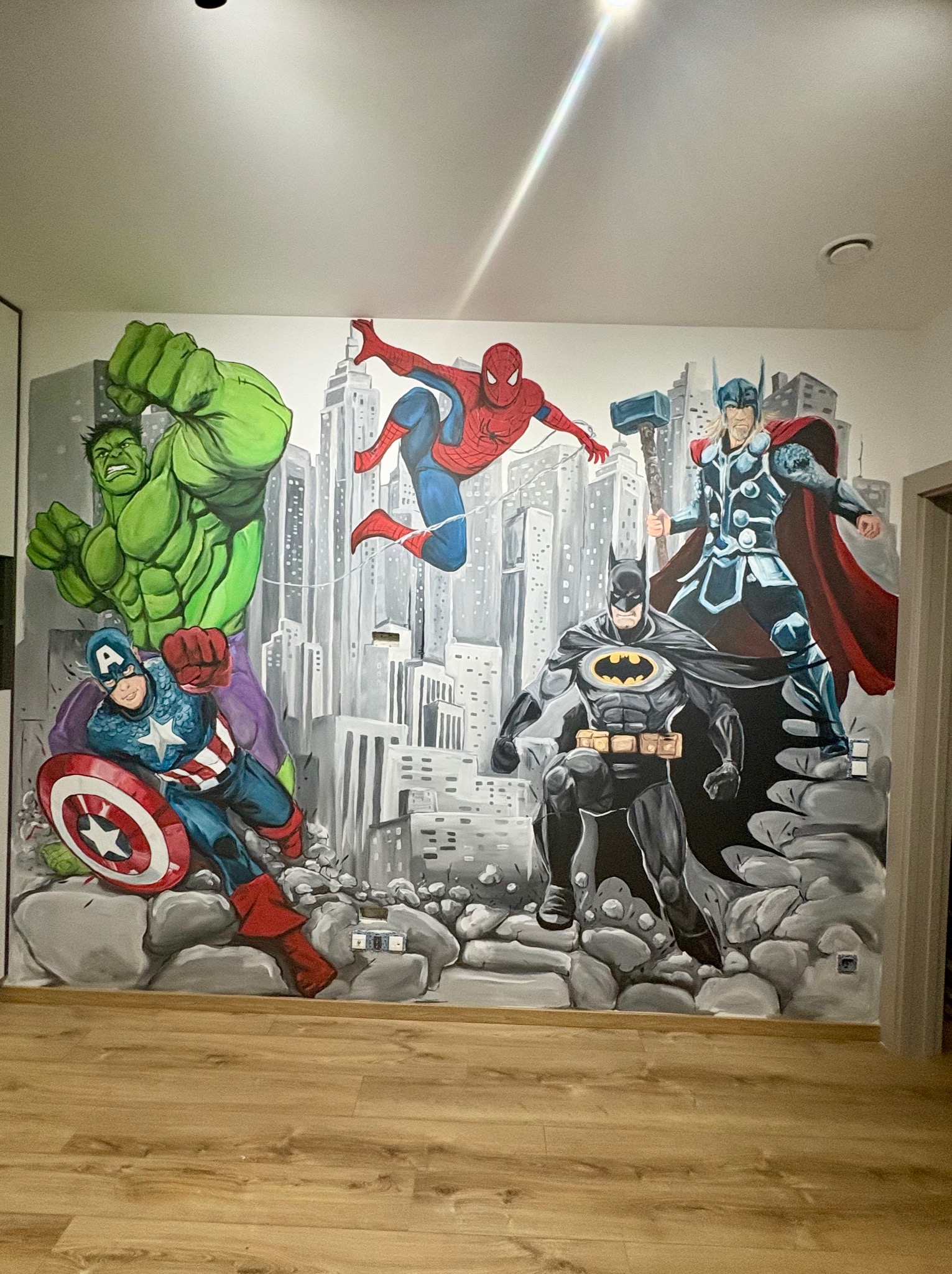 Ściana z ręcznie malowanym muralem superbohaterów (Hulk, Spider-Man, Batman, Thor, Kapitan Ameryka) na tle panoramy miasta, dół ściany imituje kamienie.