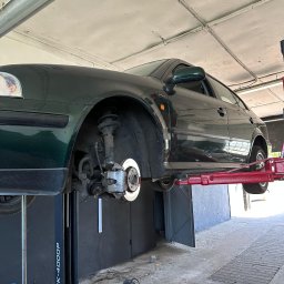 AC-AUTO - Warsztat Samochodowy - Zielony samochód na podnośniku w warsztacie, widoczne elementy zawieszenia i układu hamulcowego, narzędzia na podłodze. Model PK-4000P.