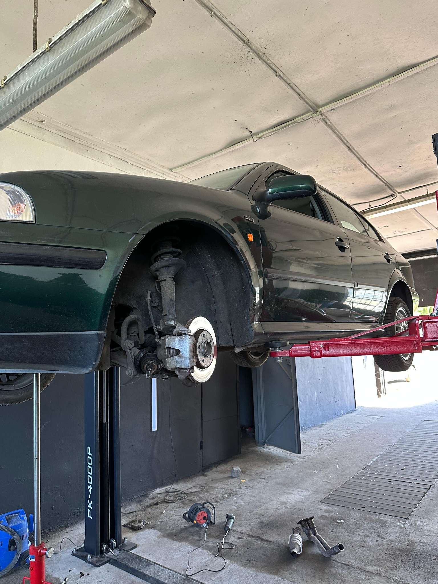 Zielony samochód na podnośniku w warsztacie, widoczne elementy zawieszenia i układu hamulcowego, narzędzia na podłodze. Model PK-4000P.